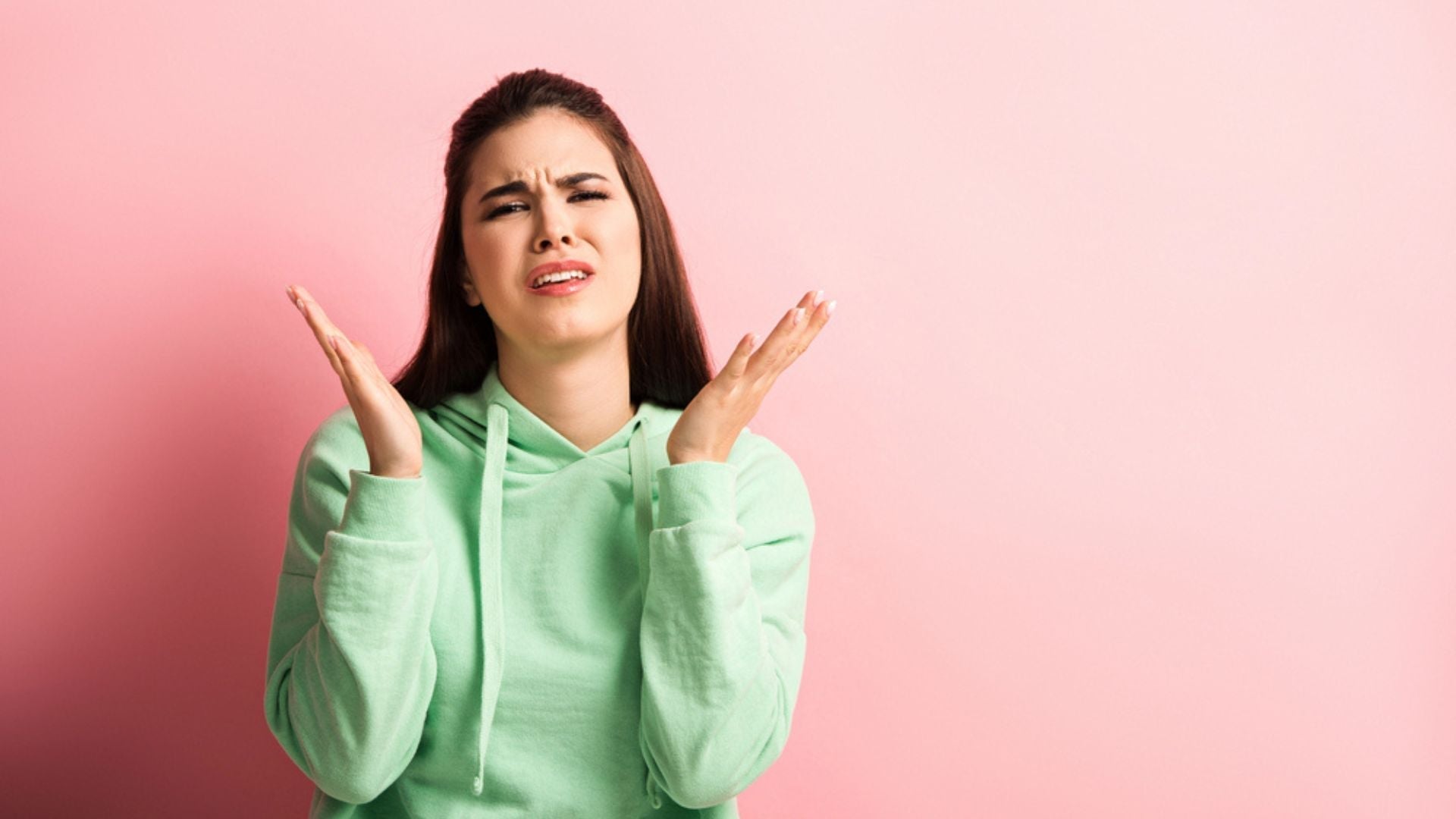 Femme déçue en pull vert, les mains levées en signe d’incompréhension, sur fond rose — expression illustrant la frustration face à un parfum qui ne tient pas.